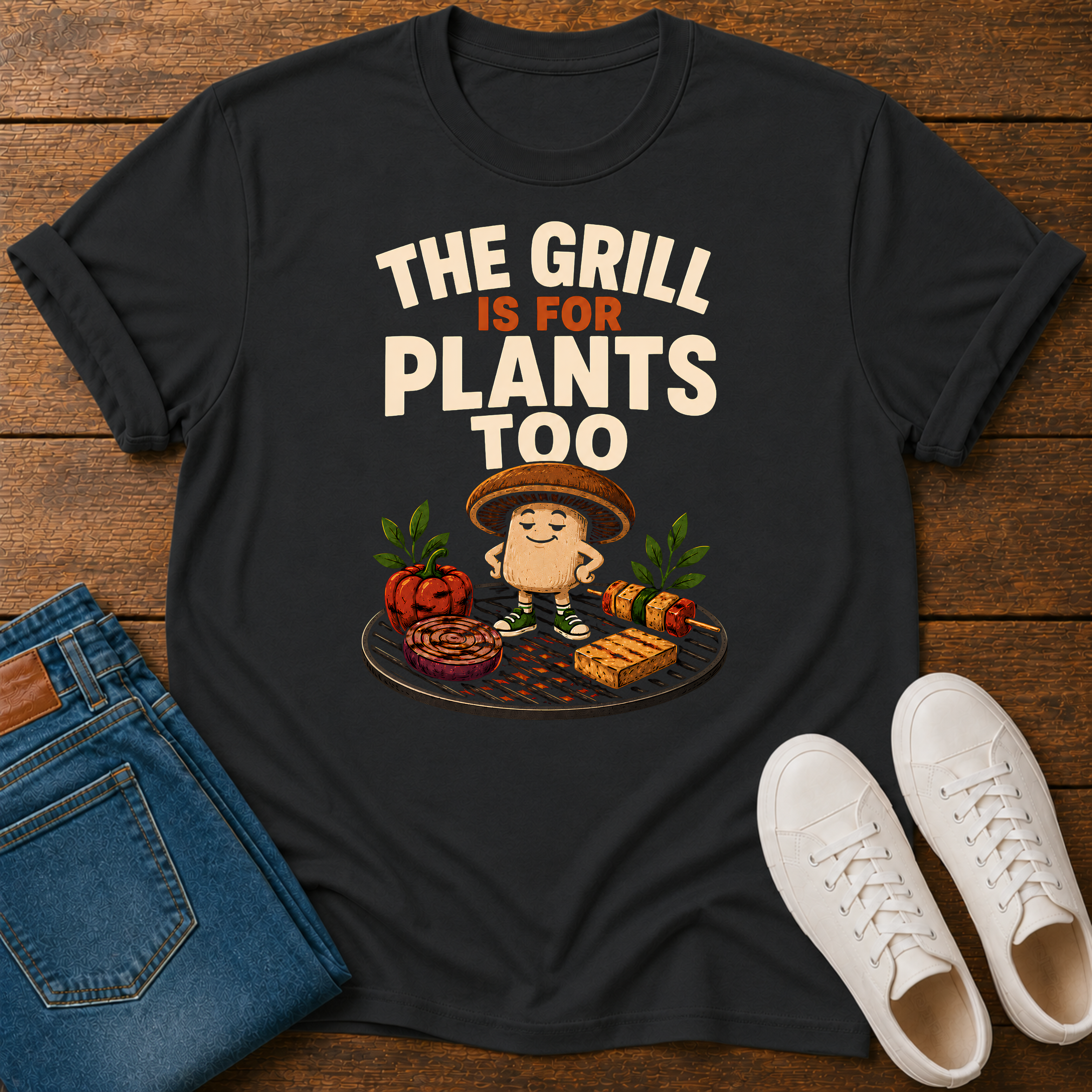 Black T-shirt with vegetables on a grill and text 'the grill is for plants too' on a wooden base with jeans and sneakers 