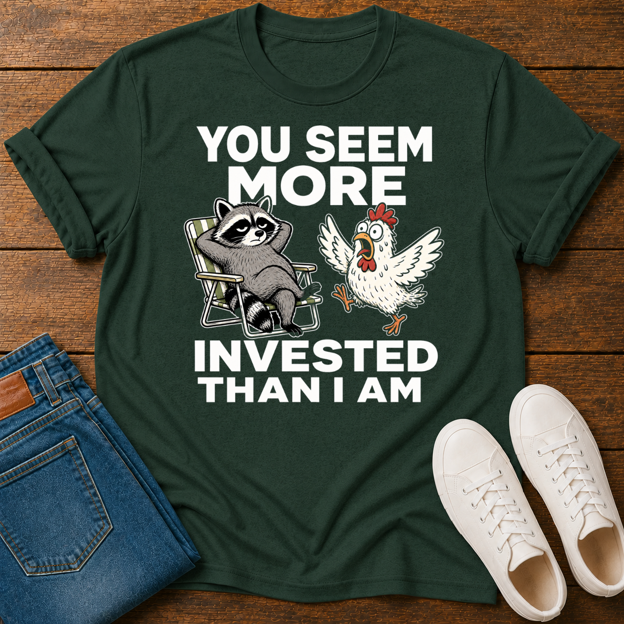Green T-Shirt with funny graphic of a raccoon being yelled at by a chicken and text 'you seem more invested than i am' on a wooden board with jeans and sneakers