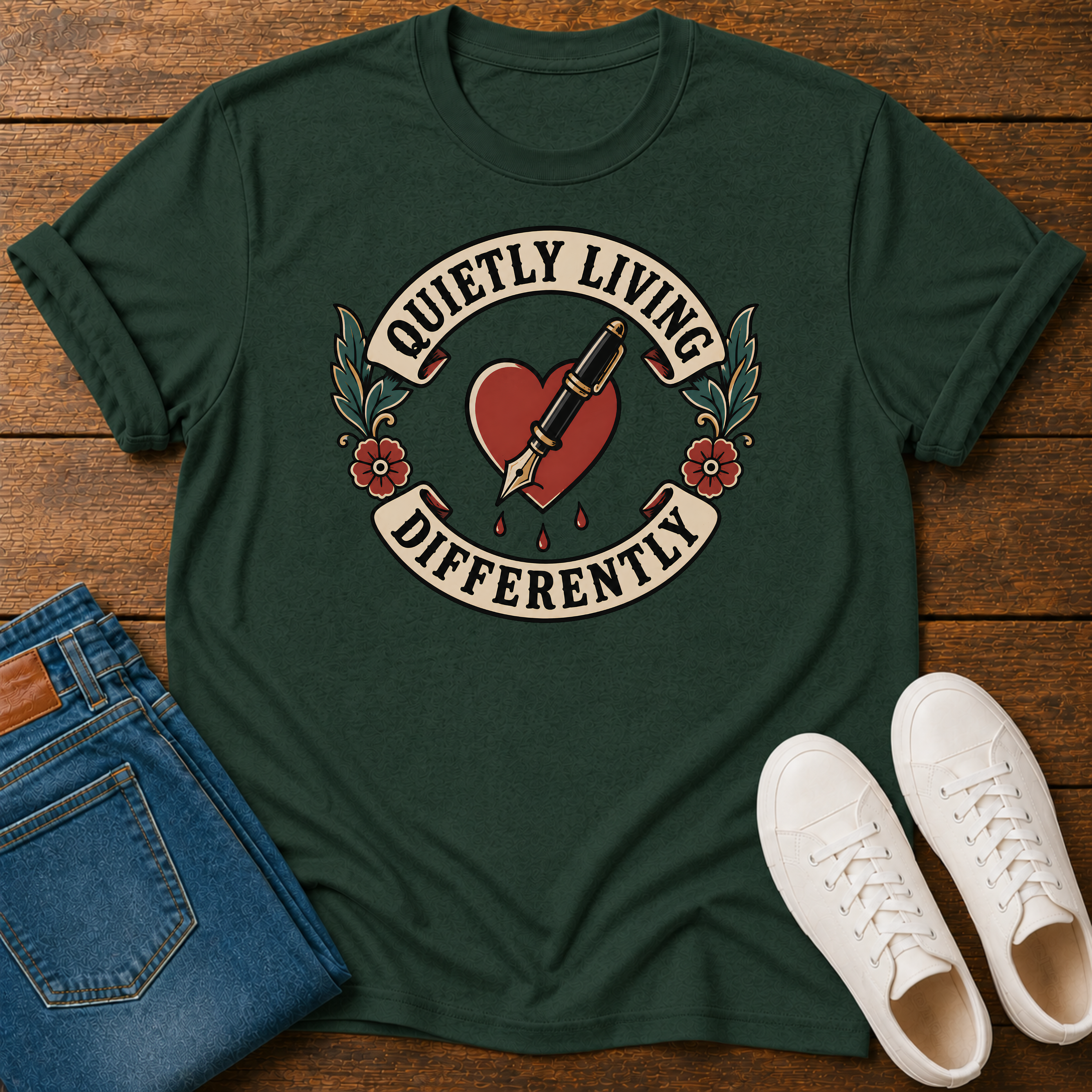 A dark green graphic t-shirt laid flat on a wooden surface, styled with folded blue jeans and white sneakers. The t-shirt features a retro tattoo-style graphic of a red heart with a fountain pen nib, surrounded by floral details and a cream-colored banner that reads 'QUIETLY LIVING DIFFERENTLY'.