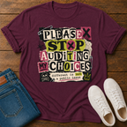 A maroon short-sleeve graphic T-shirt laid flat on a wooden surface, styled with folded blue jeans and white sneakers. The shirt has a large distressed text graphic reading 'PLEASE STOP AUDITING MY CHOICES' with small black, pink, yellow, and white blocks, plus a skull-and-crossbones detail and the phrase 'different is not a public issue' near the bottom.