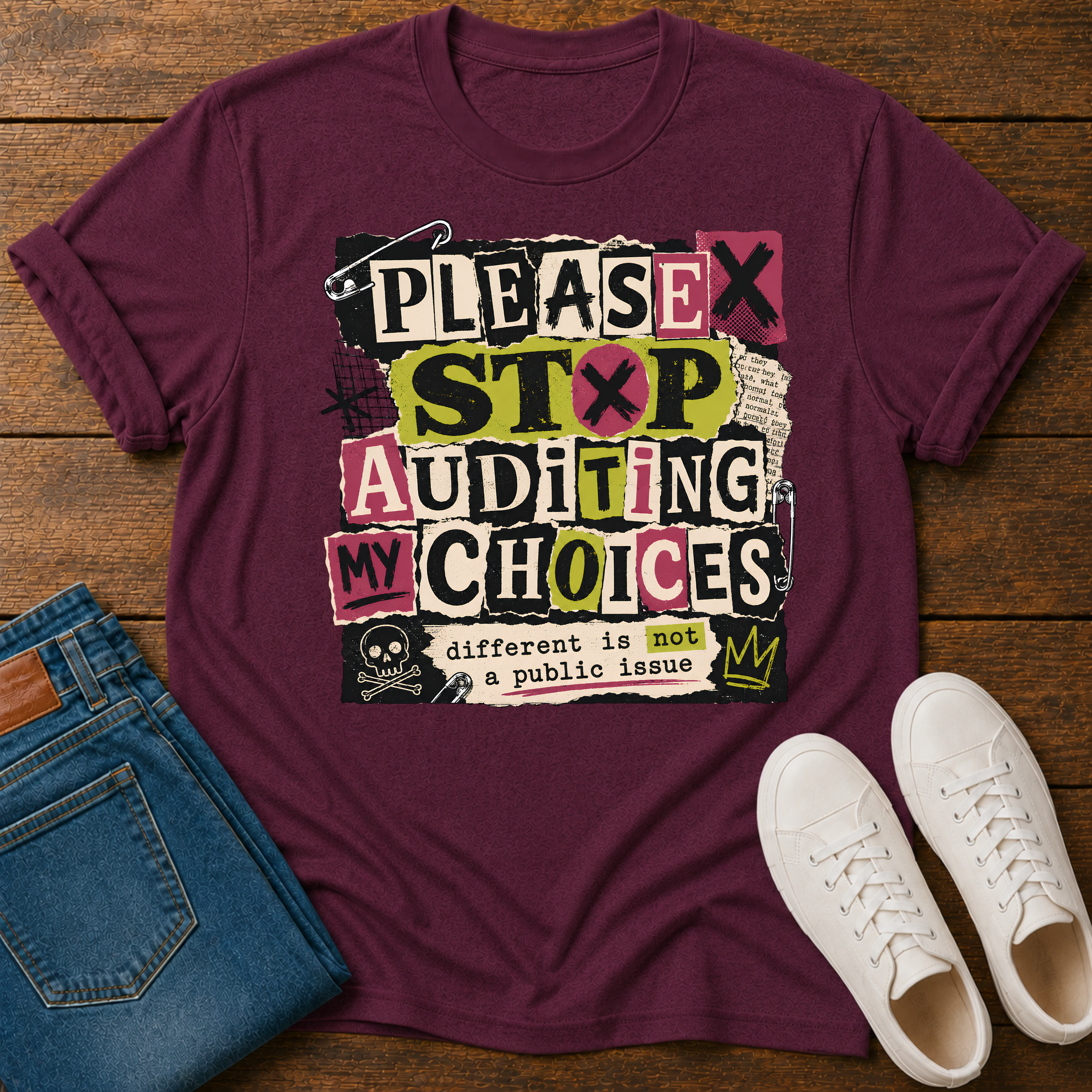 A maroon short-sleeve graphic T-shirt laid flat on a wooden surface, styled with folded blue jeans and white sneakers. The shirt has a large distressed text graphic reading 'PLEASE STOP AUDITING MY CHOICES' with small black, pink, yellow, and white blocks, plus a skull-and-crossbones detail and the phrase 'different is not a public issue' near the bottom.