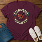 A maroon short-sleeve graphic T-shirt laid flat on a wooden surface, styled with folded blue jeans and white sneakers. The shirt has a large front graphic featuring a red heart, fountain pen, flowers, and the text 'QUIETLY LIVING DIFFERENTLY'.