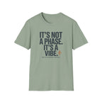 A sage green crewneck t-shirt with a dark gray text graphic reading 'IT'S NOT A PHASE. IT'S A VIBE' and a small orange stick figure icon, displayed on a clean white background.