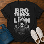 A black graphic t-shirt laid flat on a wooden surface, styled with blue jeans and white sneakers. The t-shirt features a bold white text graphic reading 'BRO THINKS HE'S A LION' above a black-and-white illustration of a man in a hoodie looking dramatic.