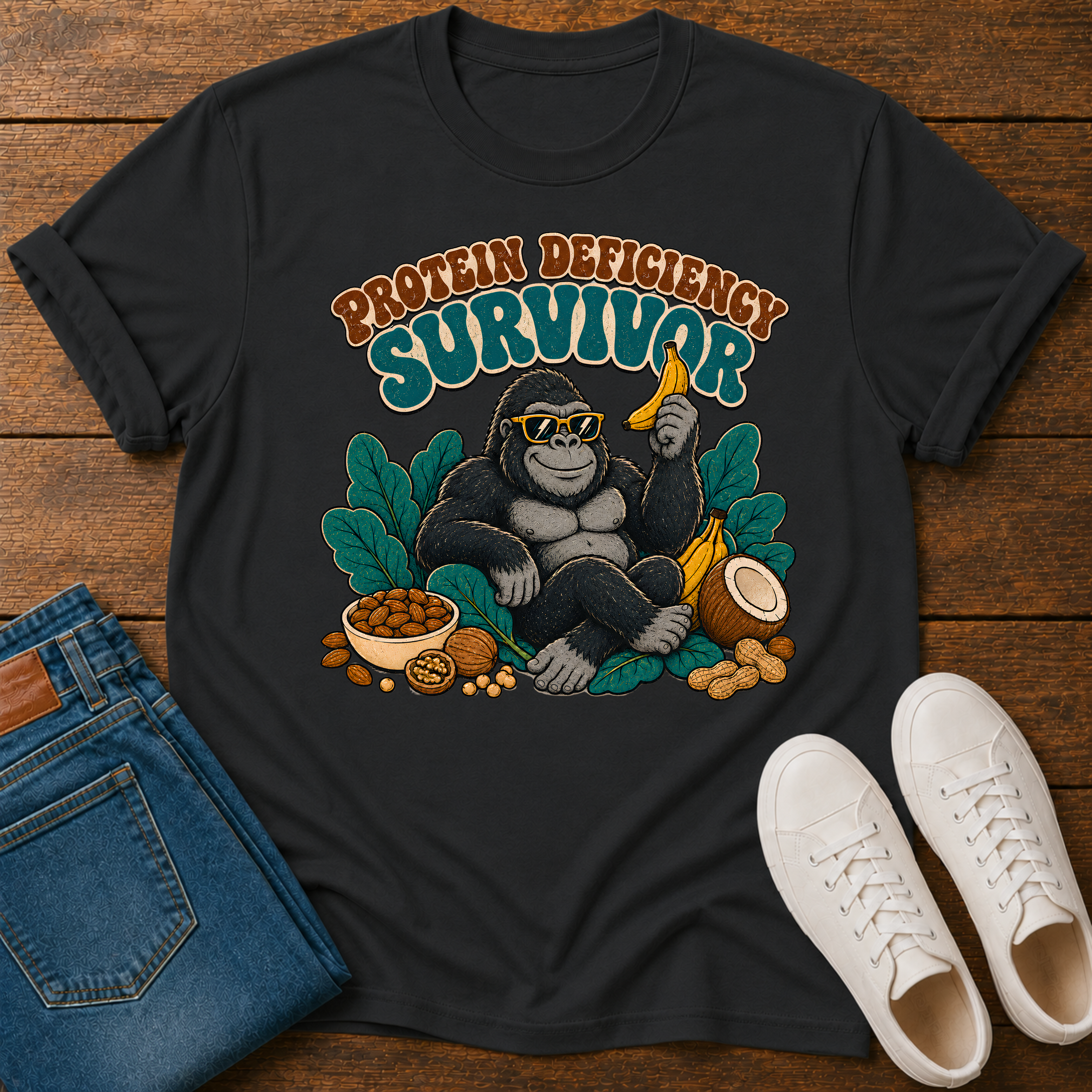 A black graphic t-shirt laid flat on a wooden surface, styled with folded blue jeans and white sneakers. The t-shirt features a green leaf crown graphic surrounding a gorilla face, with the text 'PLANT POWER' printed below in cream and red.