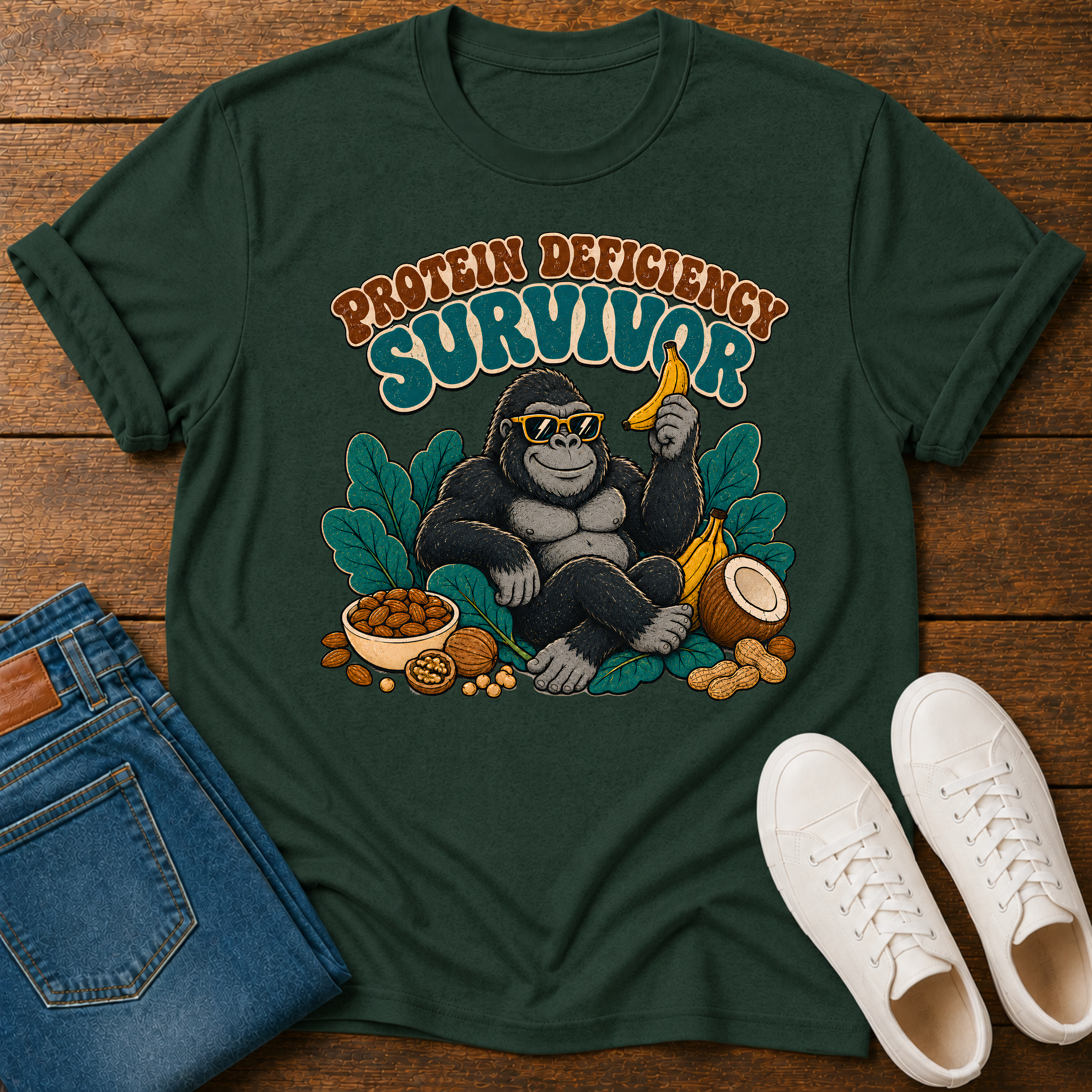 A dark green graphic t-shirt laid flat on a wooden surface, styled with folded blue jeans and white sneakers. The t-shirt features a retro-style illustration of a gorilla wearing sunglasses and holding a banana, surrounded by leaves and nuts, with the text 'PROTEIN DEFICIENCY SURVIVOR' above.