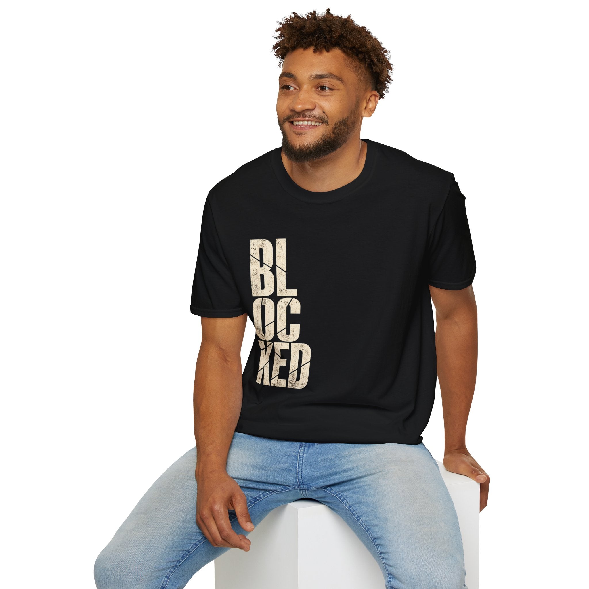 A man wearing a black crewneck t-shirt with the word 'BLOCKED' printed vertically in distressed beige block letters, paired with light blue jeans, sitting on a white block against a white background.