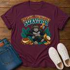 A maroon graphic t-shirt laid flat on a wooden surface, styled with blue jeans, white sneakers, and a banana. The shirt features a cartoon gorilla wearing sunglasses and holding a banana, with the text 'PROTEIN DEFICITY SURVIVOR' above it.
