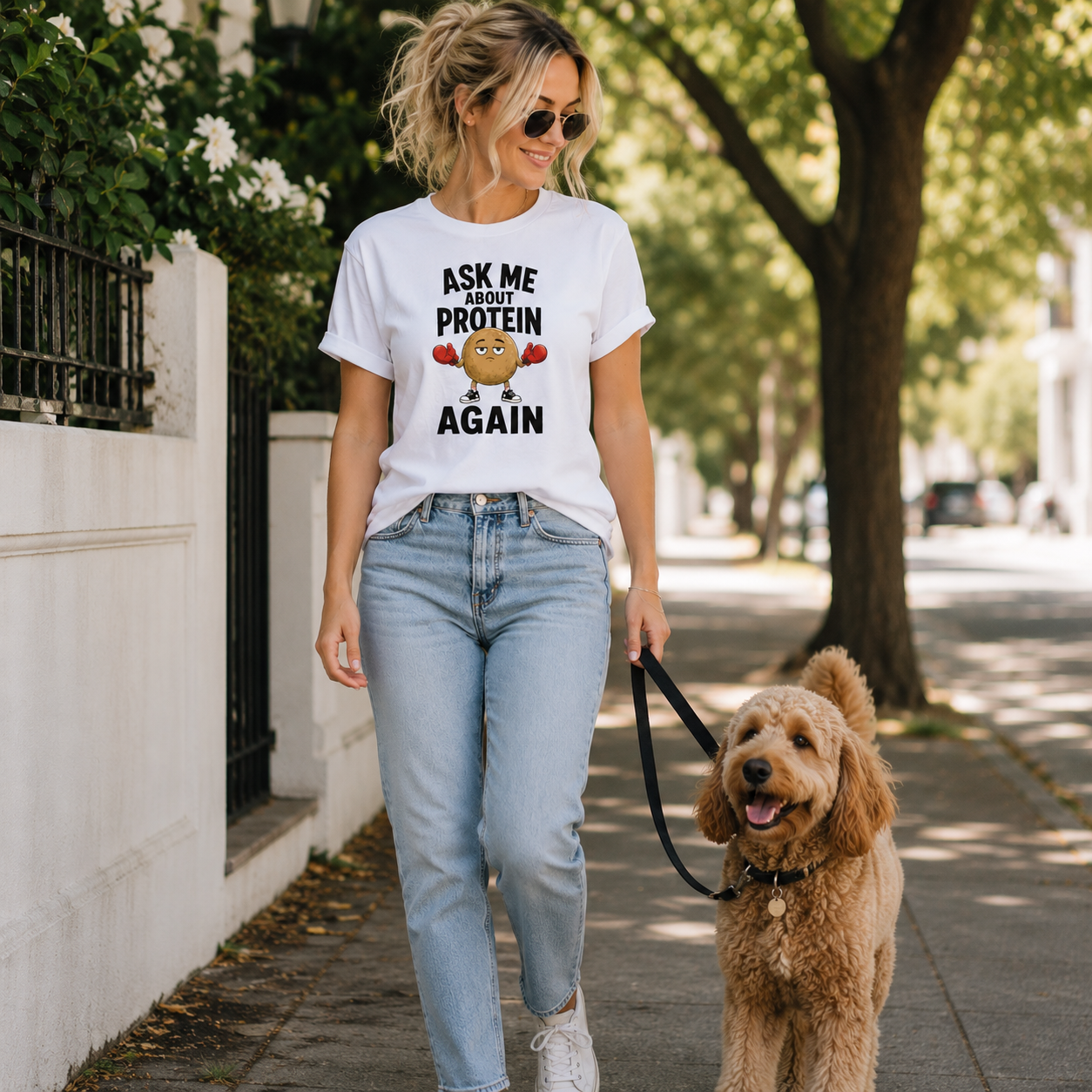A woman walking a golden dog on a sidewalk, wearing a white graphic t-shirt that reads 'ASK ME ABOUT PROTEIN AGAIN' with a chickpea character, paired with light blue jeans and white sneakers.