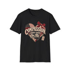 A black short-sleeve crewneck t-shirt featuring a rustic heart-shaped graphic with the text 'COMPASSION LOOKS GOOD ON ME' in a distressed cream font, decorated with a red plaid bow and ribbon details, shown against a plain white background.
