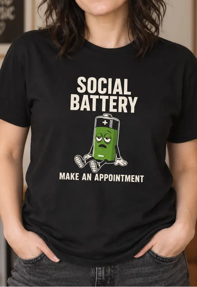 A woman wearing a black graphic t-shirt with a green cartoon battery and the text 'SOCIAL BATTERY MAKE AN APPOINTMENT'. The background is a cozy, softly lit living room with framed art, plants, and a lamp.