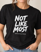 A woman wearing a black short-sleeve graphic T-shirt with the text 'NOT LIKE MOST' in large white brush-style letters and 'different by choice' underneath, paired with blue jeans against a plain light beige wall.