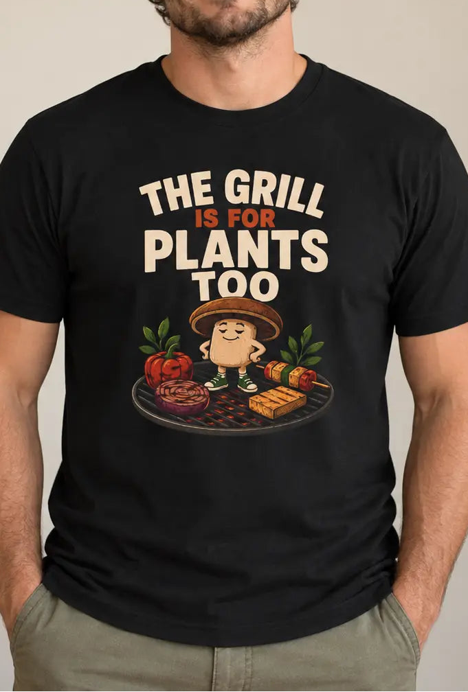A man wearing a black graphic t-shirt with the text 'THE GRILL IS FOR PLANTS TOO' and a cartoon potato grilling, standing in a room with a plant in the background.