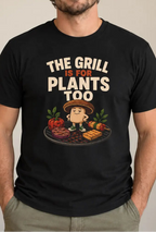 A man wearing a black graphic t-shirt with the text 'THE GRILL IS FOR PLANTS TOO' and a cartoon potato grilling, standing in a room with a plant in the background.