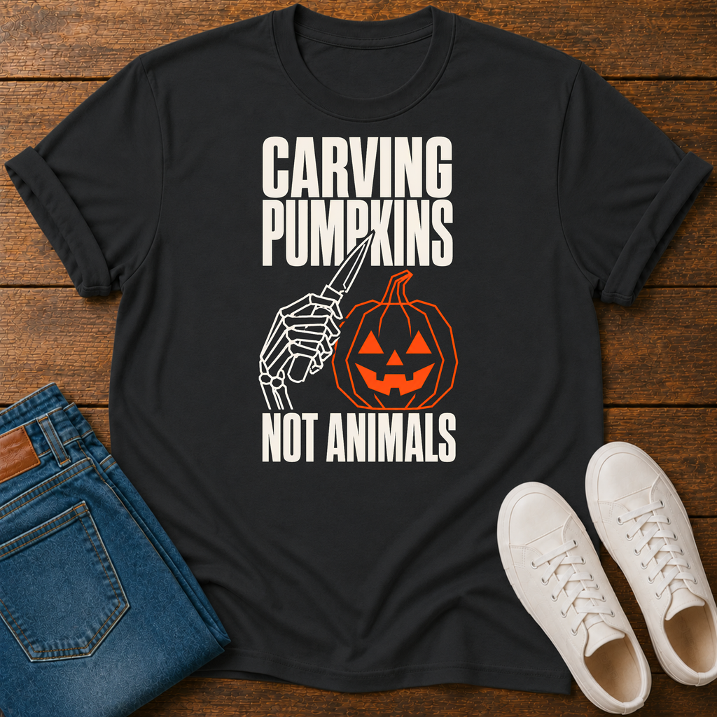 A black graphic t-shirt laid flat on a wooden surface, styled with folded blue jeans and white sneakers. The t-shirt features a graphic of a skeleton hand holding a carving knife next to a jack-o'-lantern pumpkin, with the text 'CARVING PUMPKINS NOT ANIMALS' in white and orange.