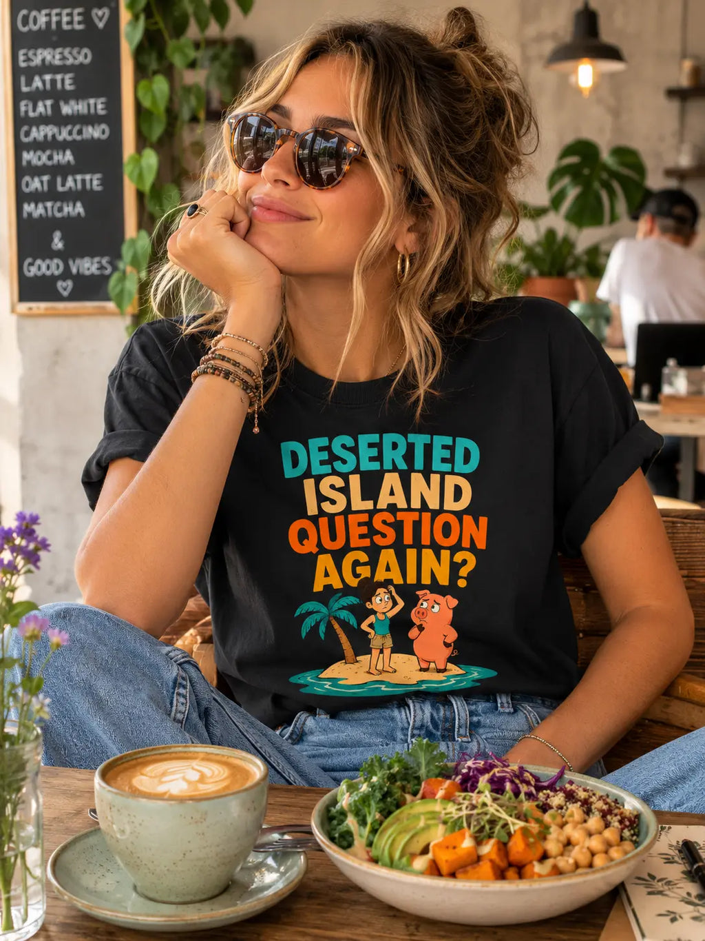 A woman wearing a black graphic t-shirt with a colorful desert island print sits in a cafe setting with a bowl of salad and a cup of coffee in front of her. Black vegan t-shirt with text 'deserted island question again?