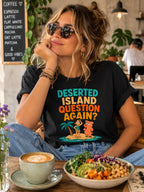 A woman wearing a black graphic t-shirt with a colorful desert island print sits in a cafe setting with a bowl of salad and a cup of coffee in front of her. Black vegan t-shirt with text 'deserted island question again?