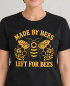 A woman wearing a black short-sleeve graphic t-shirt with a yellow bee design and the text 'MADE BY BEES LEFT FOR BEES', standing in a brightly lit indoor room with a plant in the background.