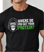 A muscular man wearing a vegan black graphic t-shirt with the text 'WHERE DO YOU GET YOUR PROTEIN?' in white and green next to an illustrated gorilla head. The background is a gym with dumbbells and a motivational poster.