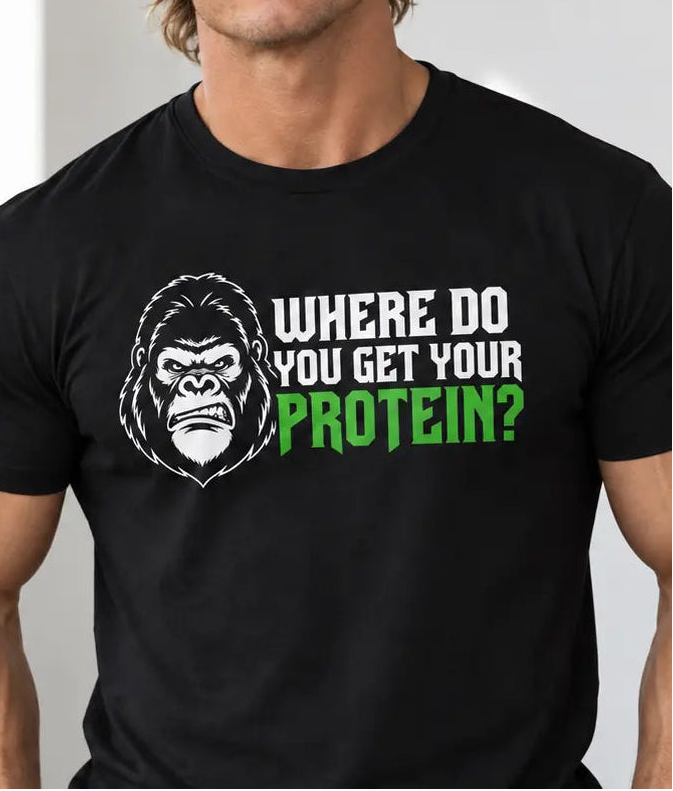 A muscular man wearing a vegan black graphic t-shirt with the text 'WHERE DO YOU GET YOUR PROTEIN?' in white and green next to an illustrated gorilla head. The background is a gym with dumbbells and a motivational poster.