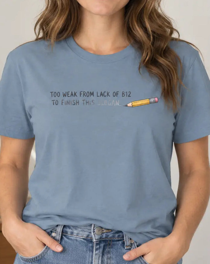 A woman wearing a dusty blue graphic t-shirt with the text 'TOO WEAK FROM LACK OF B12 TO FINISH THIS SLOGAN' and a small pencil illustration, paired with blue jeans in a bright indoor setting.