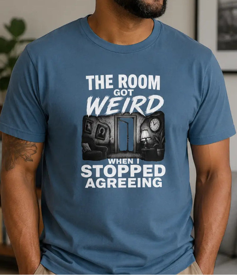 A man wearing a blue graphic t-shirt with the text 'THE ROOM GOT WEIRD WHEN I STOPPED AGREING' and a graphic of two robots, standing in a living room setting.