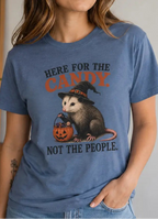 A woman wearing a halloween blue graphic t-shirt with a cartoon opossum wearing a witch hat and holding a jack-o'-lantern, with the text 'HERE FOR THE CANDY NOT THE PEOPLE.' She is wearing light wash blue jeans in an indoor setting.
