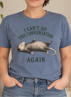 A woman wearing a slate blue graphic t-shirt with a sleeping sloth illustration and the text 'I CAN'T DO THIS CONVERSATION AGAIN'.