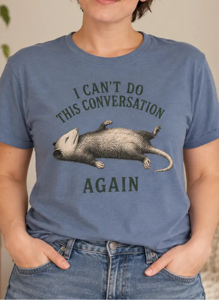 A woman wearing a slate blue graphic t-shirt with a sleeping sloth illustration and the text 'I CAN'T DO THIS CONVERSATION AGAIN'.