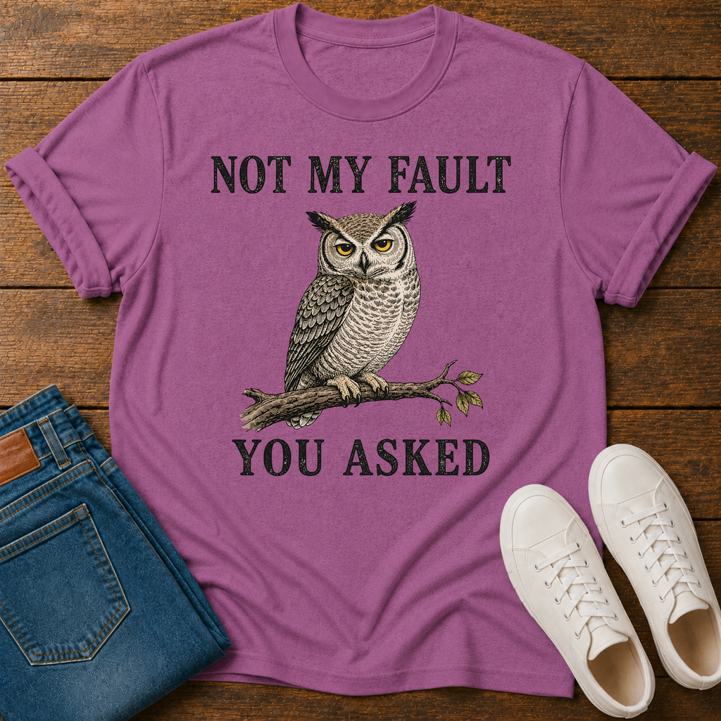 A bright pink  graphic t-shirt laid flat on a wooden surface, styled with blue jeans, white sneakers. The t-shirt features a graphic of a owl perched on a branch with the text 'NOT MY FAULT YOU ASKED' in black.