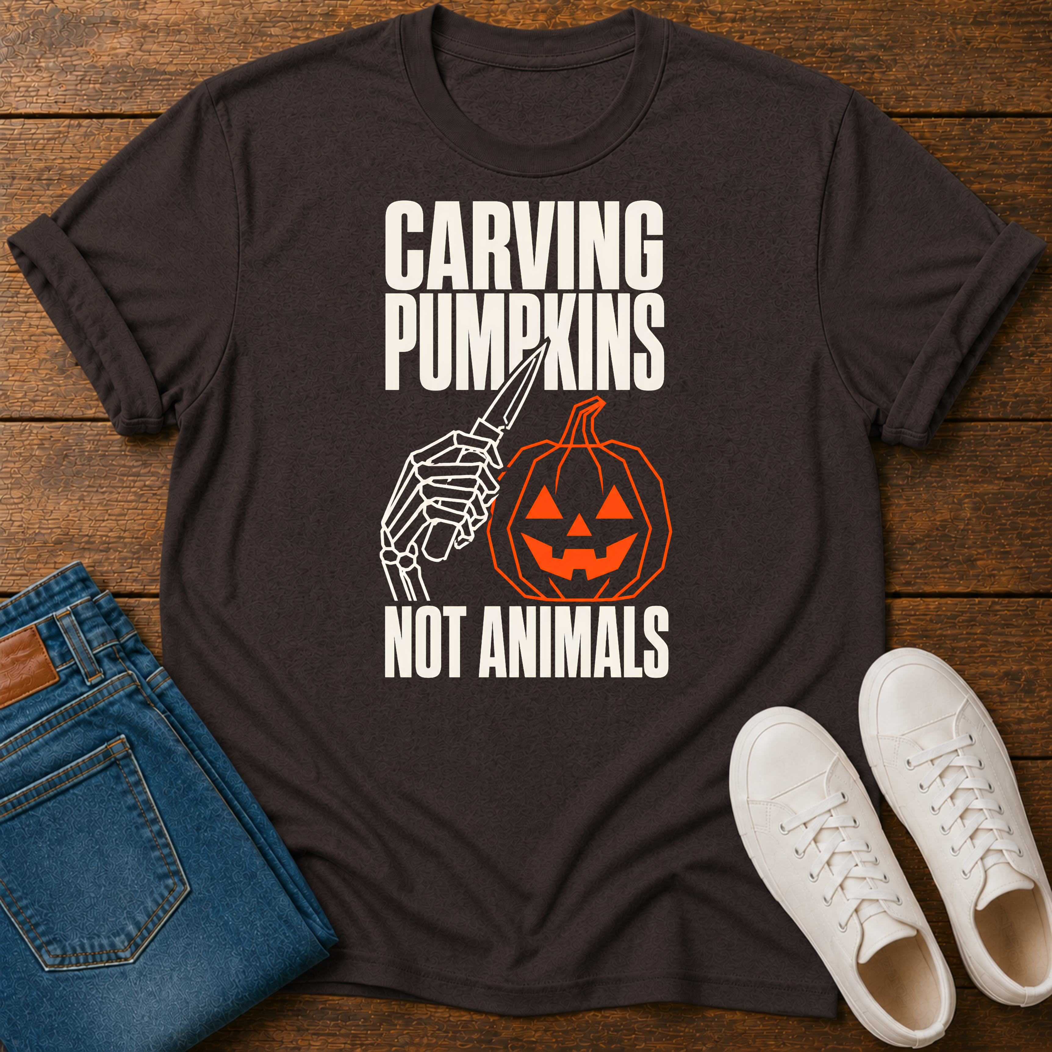 A dark brown graphic t-shirt laid flat on a wooden surface, styled with blue jeans and white sneakers. The shirt features a Halloween-themed graphic with the text 'CARVING PUMPKINS NOT ANIMALS' in white, showing a skeleton hand holding a knife next to a carved orange pumpkin.