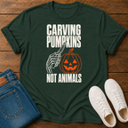 A green graphic t-shirt laid flat on a wooden surface, styled with blue jeans and white sneakers. The shirt features a Halloween-themed graphic with the text 'CARVING PUMPKINS NOT ANIMALS' in white, showing a skeleton hand holding a knife next to a carved orange pumpkin.