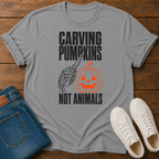 A sports gray graphic t-shirt laid flat on a wooden surface, styled with folded blue jeans and white sneakers. The t-shirt features a graphic of a skeleton hand carving a jack-o'-lantern with the text 'CARVING PUMPKINS NOT ANIMALS' in black and orange.