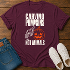 A maroon graphic t-shirt laid flat on a wooden surface, styled with blue jeans and white sneakers. The shirt features a Halloween-themed graphic with the text 'CARVING PUMPKINS NOT ANIMALS' in white, showing a skeleton hand holding a knife next to a carved orange pumpkin.