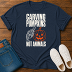 A navy blue graphic t-shirt laid flat on a wooden surface, styled with folded blue jeans and white sneakers. The t-shirt features a Halloween-themed graphic with the text 'CARVING PUMPKINS NOT ANIMALS' in white, alongside a skeleton hand holding a knife next to a carved orange pumpkin.