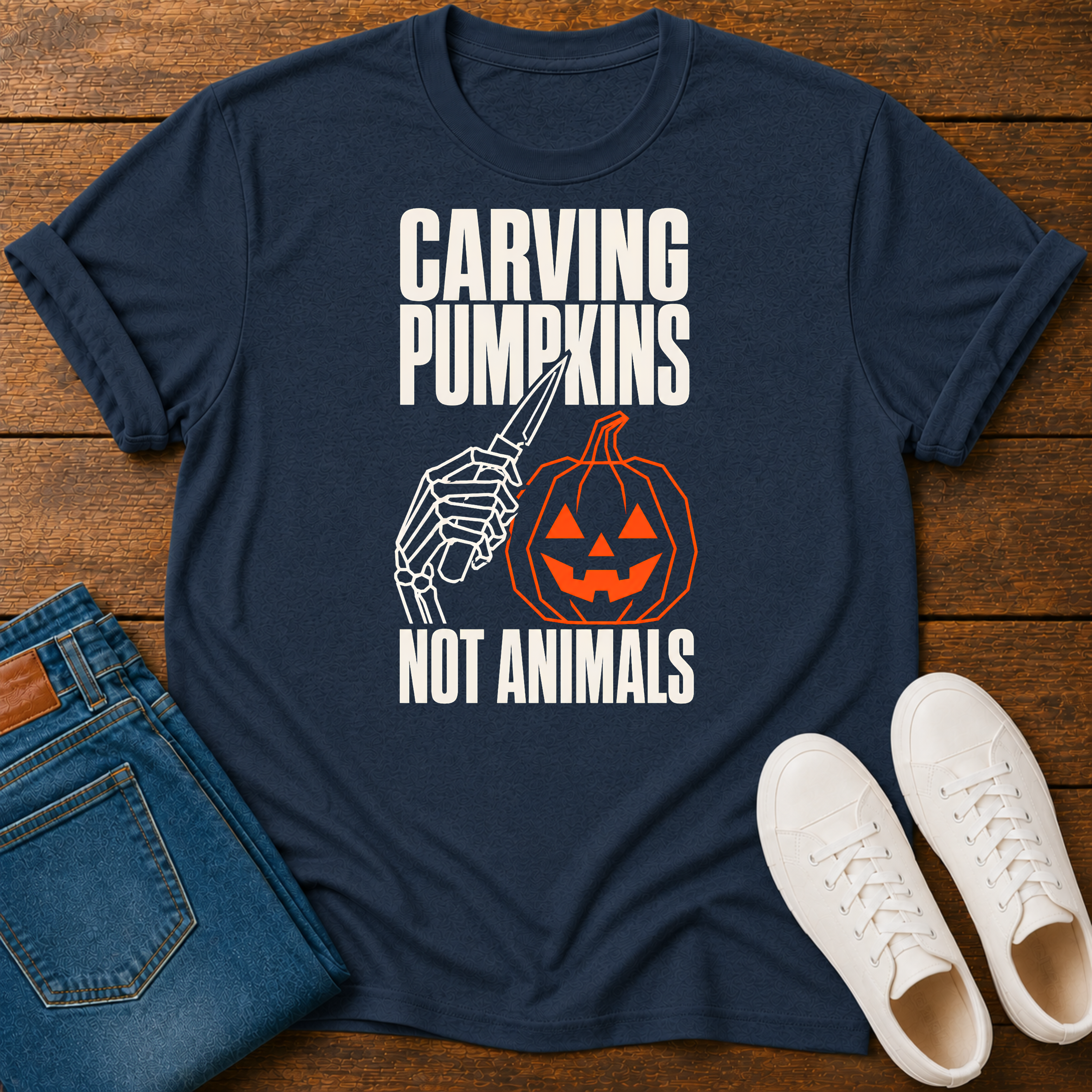 A navy blue graphic t-shirt laid flat on a wooden surface, styled with folded blue jeans and white sneakers. The t-shirt features a Halloween-themed graphic with the text 'CARVING PUMPKINS NOT ANIMALS' in white, alongside a skeleton hand holding a knife next to a carved orange pumpkin.