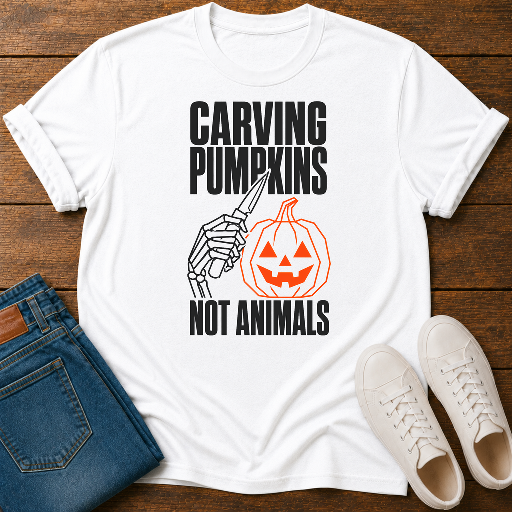 A white graphic t-shirt laid flat on a dark wood background, styled with folded blue jeans and a pair of white sneakers. The t-shirt features a black and orange graphic of a skeleton hand carving a jack-o'-lantern with the text 'CARVING PUMPKINS NOT ANIMALS'.