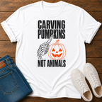 A white graphic t-shirt laid flat on a dark wood background, styled with folded blue jeans and a pair of white sneakers. The t-shirt features a black and orange graphic of a skeleton hand carving a jack-o'-lantern with the text 'CARVING PUMPKINS NOT ANIMALS'.