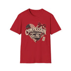 A red short-sleeve crewneck t-shirt featuring a distressed graphic on the front. The graphic shows a heart made of American flag and plaid patterns with a red bow, and the text 'COMPASSION LOOKS GOOD On Me' in a vintage cream font.
