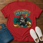 A red graphic t-shirt featuring a gorilla sitting with banana, nuts, and the text 'PROTEIN DEFICITY SURVIVOR'. The shirt is styled in a flat-lay on a wooden background alongside blue jeans, white sneakers, and a small bowl of peanut butter.