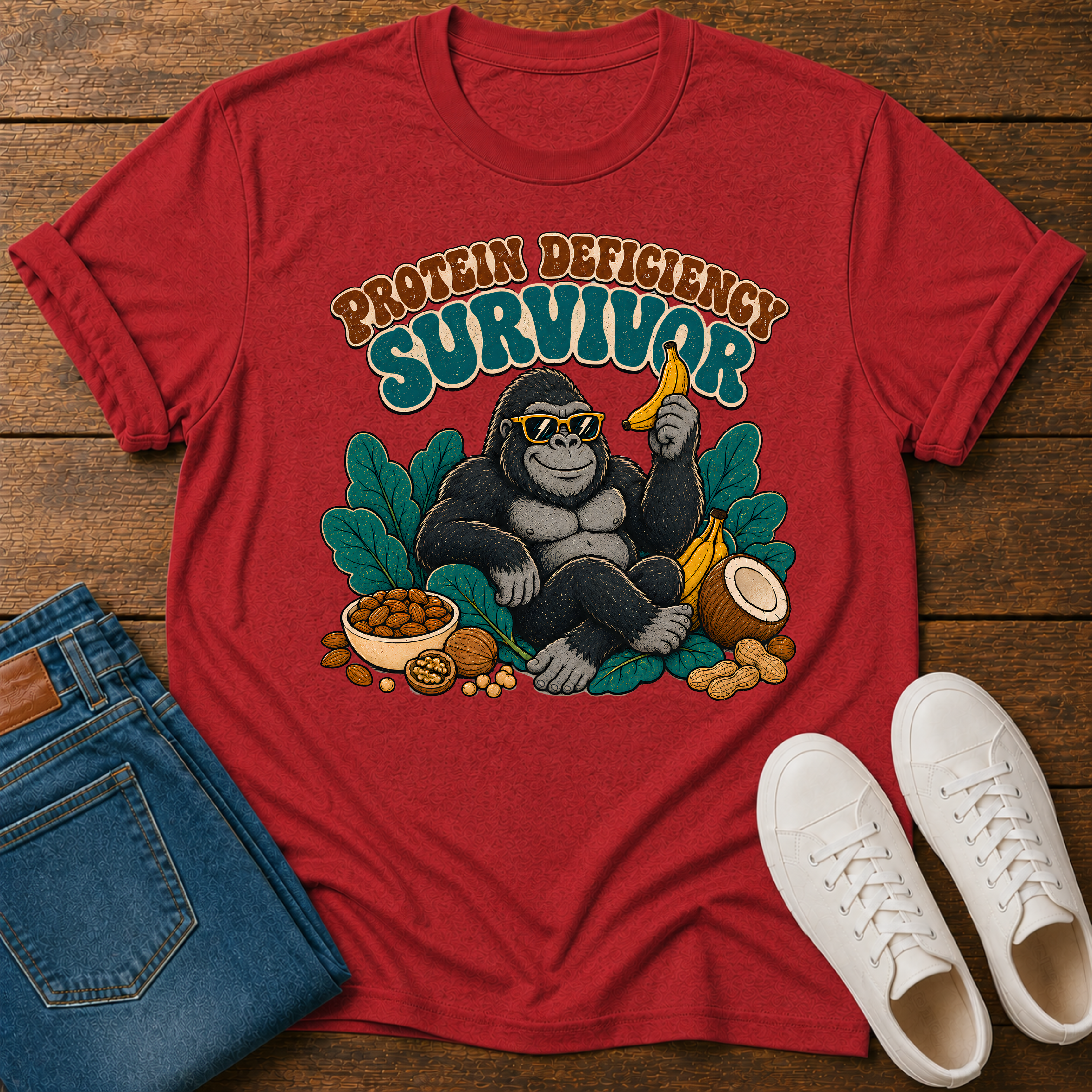 A red graphic t-shirt featuring a gorilla sitting with banana, nuts, and the text 'PROTEIN DEFICITY SURVIVOR'. The shirt is styled in a flat-lay on a wooden background alongside blue jeans, white sneakers, and a small bowl of peanut butter.