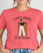 Woman wearing a coral pink shirt with graphic of a tall woman and text 'i don't shrink to fit rooms'