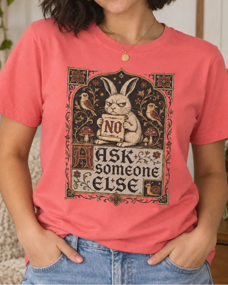 A woman wearing a coral pink graphic t-shirt with a vintage-style illustration of a rabbit and the text 'ASK SOMEONE ELSE NO'. She is posing in a cozy, decorated room with plants and a framed quote on the wall.
