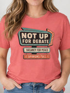 A woman wearing a coral pink graphic t-shirt with a retro marquee design that reads 'NOT UP FOR DEBATE', 'VACANCY FOR PEACE', and 'OPINIONS FULL'. She is standing in a brightly lit room with a framed sign on the wall and a plant in the background.