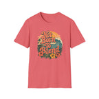 A coral pink short-sleeve t-shirt with a retro-style graphic on the chest reading 'Not Born To Blend' in orange lettering, surrounded by a wave, sunset, and tropical flowers.