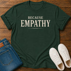 A dark forest green t-shirt with the word 'EMPATHY' printed in large cream-colored letters, styled in a flat lay on a wooden background alongside folded blue jeans and a pair of white sneakers.