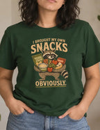 A woman wearing a dark green graphic t-shirt with a cartoon raccoon holding snacks and the text 'I BROUGHT MY OWN SNACKS OBVIOUSLY.' in a cozy indoor setting with plants and home decor.