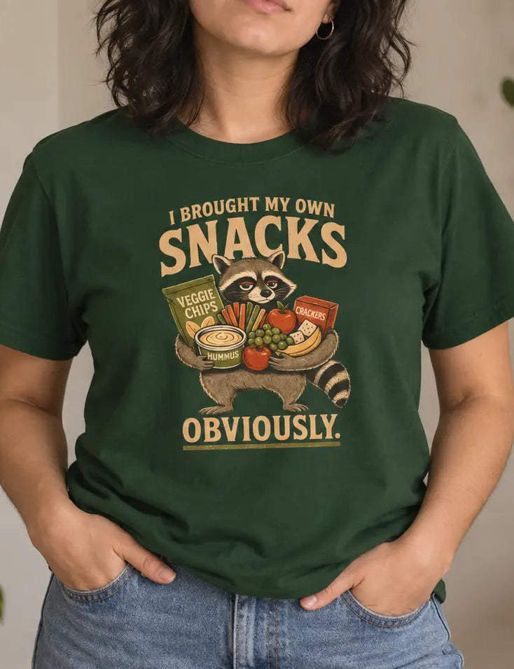 A woman wearing a dark green graphic t-shirt with a cartoon raccoon holding snacks and the text 'I BROUGHT MY OWN SNACKS OBVIOUSLY.' in a cozy indoor setting with plants and home decor.