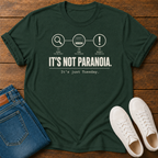 A green short-sleeve graphic T-shirt laid flat on a wooden surface, styled with folded blue jeans and white sneakers.  text 'it's not paranoia it's a tuesday'