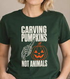 A woman wearing a dark green graphic t-shirt with the text 'CARVING PUMPKINS NOT ANIMALS' and a skeleton hand carving a pumpkin illustration, posed in a cozy indoor setting.